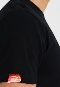 Černé bavlněné tričko s kulatým výstřihem, s viditelnou červenou a bílou cedulkou "SuperDry" na levém rukávu. Hladká textura, přiléhavý střih.
