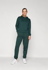 Sweat à capuche vert foncé avec une coupe décontractée, pantalon de jogging assorti et baskets blanches. Le matériau semble doux, avec des manches longues et des poignets côtelés.