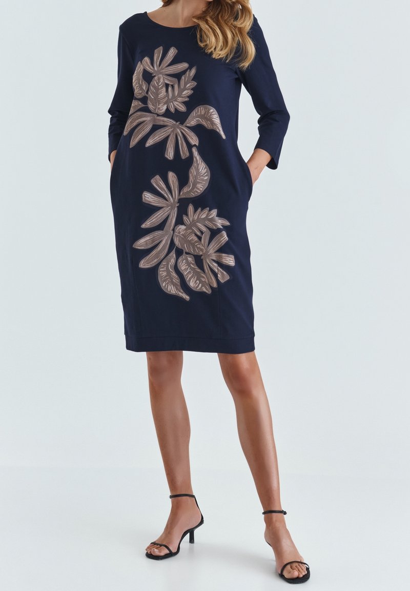 Femme portant une robe bleu marine jusqu'aux genoux avec des manches trois-quarts et des motifs de feuilles beiges, assortie à des sandales à talons noirs.