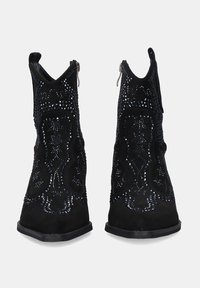 Botines de ante negros con puntera puntiaguda, tachuelas decorativas, intrincados patrones de costura y cierres de cremallera lateral, mostrados de frente.