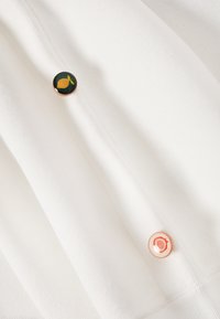 Tissu blanc avec deux boutons circulaires, l'un représentant un citron jaune sur fond vert foncé, l'autre une grenade rose sur fond rose clair.