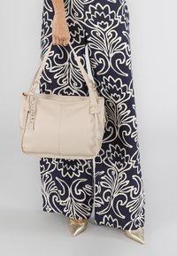 Borsa a mano in pelle beige con due manici corti, hardware dorato e finitura texturizzata. La borsa è mostrata abbinata a pantaloni con fantasia.