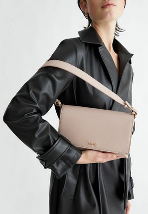 Sac en cuir rose clair porté en bandoulière, avec une surface texturée, un logo doré et une bandoulière ajustable, porté contre un manteau en cuir noir.