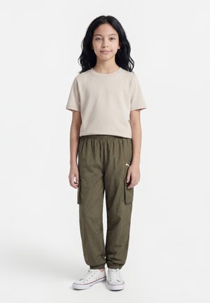 Dievča s dlhými čiernymi vlasmi, oblečené do béžového trička, olivovo zelených nohavíc cargo a bielych tenisiek, stojace na jednoduchom bielom pozadí.