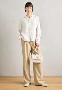 Linnen knoopshirt in gebroken wit, gecombineerd met losse beige broeken. Houdt een zachte crèmekleurige tas vast met een gouden hardware detail. Platte schoenen.