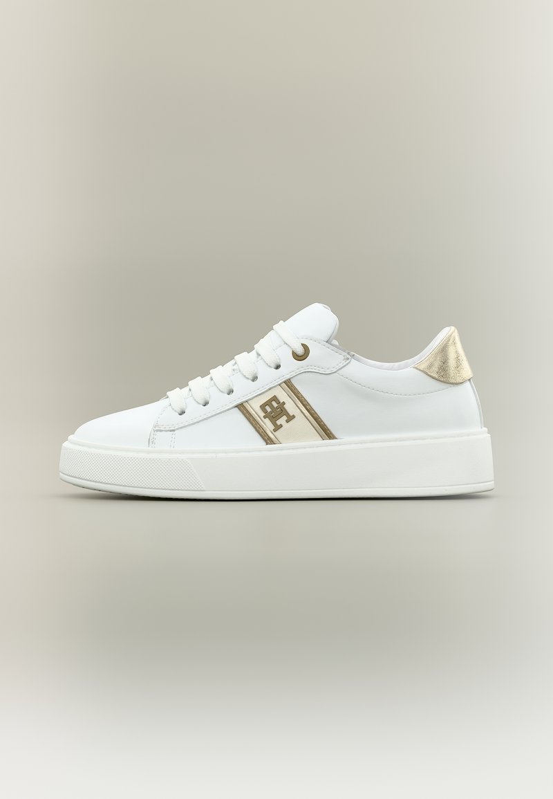 Λευκό sneaker χαμηλού πάτου με χρυσή λεπτομέρεια στη φτέρνα, χρυσό λογότυπο και ρίγα, καθώς και χοντρή λευκή σόλα, εμφανίζεται σε προφίλ πλευρικής όψης σε ουδέτερο φόντο.