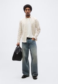 Homme portant une chemise blanche à rayures, un t-shirt blanc, un jean bleu ample, des chaussures noires, tenant un sac de sport noir de la main gauche dans une pose neutre.