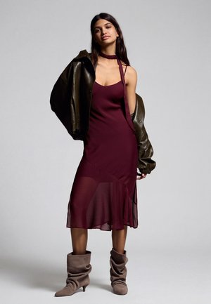 Nő bordó ujjatlan ruhában, vállán bőrkabáttal, és laza taupe bokacsizmában, egyszerű háttér előtt áll.