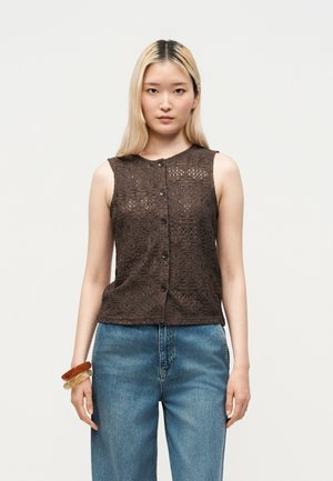 Jonge vrouw met steil blond haar die een mouwloze donkerbruine kanten top met knopen en een blauwe spijkerbroek draagt, geaccentueerd met armbanden aan de linkerpols.