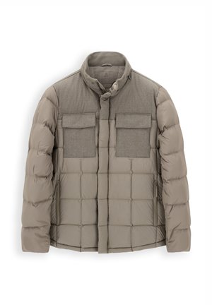Homme portant une veste matelassée gris clair, une chemise beige, un jean bleu foncé, une ceinture marron et des baskets beiges, debout devant un fond uni.