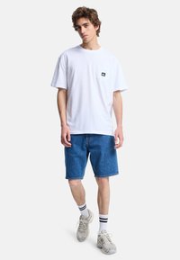Hvid kortærmet t-shirt med en venstre brystlomme, parret med blå denimshorts. Fodtøjet er beige sneakers med grå accenter.