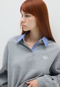 Sudadera gris con un cuello a rayas en azul y blanco, que presenta un pequeño corazón bordado y un logo con letras en el pecho. Textura suave.