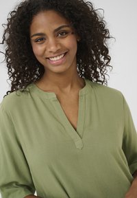 Blouse vert olive avec un décolleté en V et des manches courtes, faite d'un tissu doux avec une texture lisse, présentant un design simple et épuré.