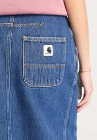Fustă din denim albastru cu un buzunar pe spate, având o patch cu logo-ul Carhartt alb. Țesătura are o textură clasică și detalii de cusătură vizibile.