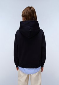Sudadera negra con capucha, de textura suave, con un bolsillo tipo canguro y un dobladillo acanalado, superpuesta a una camisa azul claro, combinada con pantalones beiges.