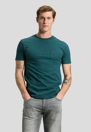 Junger Mann mit kurzen Haaren, der ein grünes kurzärmeliges T-Shirt und hellgraue Jeans trägt, steht mit den Händen in den Taschen vor einem schlichten Hintergrund.