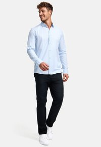 Camisa de botones azul claro con mangas largas, combinada con pantalones negros y zapatillas blancas. La camisa presenta un cuello clásico y detalles de botones.