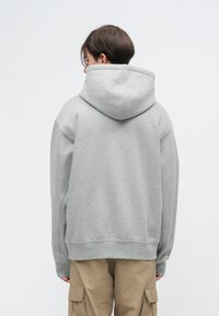 Sivi preveliki hoodie od mekog materijala, s velikom kapuljačom i rebrastim manžetama. Nosite ga uz bež cargo hlače, viđeno s leđa.