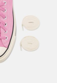 Różowy zamszowy sneaker o fakturowanej powierzchni, z białym gumowym noskiem i srebrnymi oczkami. Obok buta leżą dwie zwinięte kremowe sznurowadła.