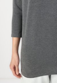 Grå långärmad topp av mjukt material, med en avslappnad passform och rund fåll, som sträcker sig till mitten av låret på bäraren.