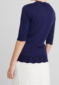 Námořnicky modrý pletený svetr s krátkými rukávy, vroubkovaným výstřihem a krajkovým vzorem na rukávech a lemu. Hladká textura, přiléhavý střih.
