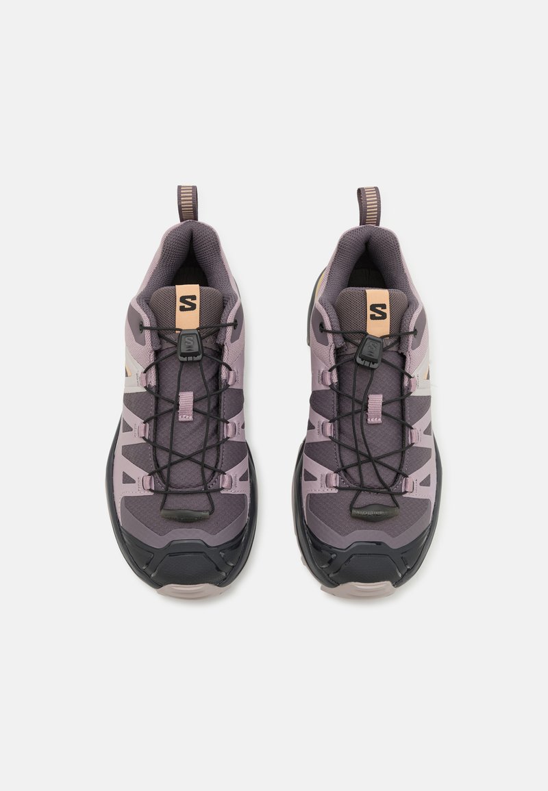 Zapatillas deportivas moradas con parte superior de malla, suela de goma negra y cordones elásticos. Presentan una pestaña en bucle y acentos claros contrastantes en los lados.