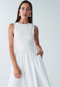 Robe blanche avec un corsage froncé et une jupe évasée. Le tissu a une texture lisse, et la robe possède des poches et un design sans manches.