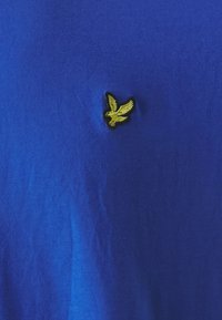 T-shirt in cotone blu con una texture liscia, presenta un piccolo logo ricamato d'oro a forma di aquila sul petto. Design semplice con collo rotondo.
