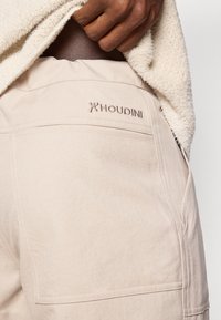 Beežid Houdini lühikesed püksid, millel on sile tekstuur, elastne vöökoht ja diskreetne tagatasku õmblus. Logo on tikandina tagaküljel.