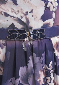 Námořnický modrý pásek s lesklou sponou ve tvaru listu přes fialovou květinovou plisovanou látku šatů.