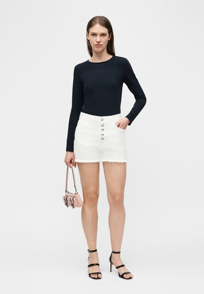 Γυναίκα με μαύρο μακρυμάνικο τοπ, λευκά σορτς με κουμπιά, μαύρα πέδιλα με λουριά, κρατώντας μια μικρή ροζ τσάντα χειρός, στέκεται μπροστά σε λευκό φόντο.