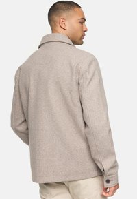 Veste en laine beige avec un col à revers, coupe droite et détails de poignets à boutons. Texture douce, design minimaliste et sans motifs visibles.