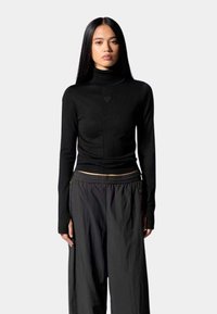 Un pull à col roulé noir au design ajusté, fabriqué en tissu doux, avec un petit détail de logo sur la poitrine et des manches longues. Porté avec un pantalon noir ample.