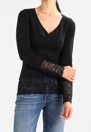 Femme portant un haut noir à manches longues avec des poignets et un ourlet en dentelle, associé à un jean bleu, debout devant un fond blanc.