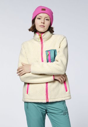 Young woman wearing a cream fleece jacket with pink zippers and a teal pocket, teal pants, and a bright pink knit hat, standing with arms crossed.