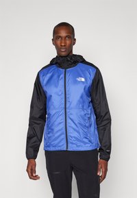 Giacca leggera blu e nera con chiusura a zip, cappuccio regolabile e tasche laterali. Presenta un logo sul petto e una superficie liscia.