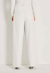 Frau trägt hochgeschnittene, weit geschnittene weiße Hosen mit Gürtelschlaufen, Gesäßtasche und facettierten hellgrauen High Heels.