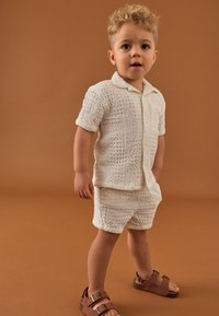 Cămașă texturată albă cu mâneci scurte și pantaloni scurți asortați, cu un model subtil. Sandale maro cu două curele cu cataramă. Fundal maro.