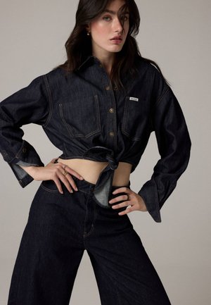 Le mannequin pose les mains sur les hanches, portant une chemise en denim foncé nouée à la taille et un jean large assorti.