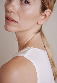 Hvid ærmeløs top med rund halsudskæring, blød tekstur og minimal syning. Modellen har langt, glat hår, som er delvist synligt.