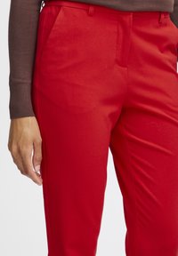 Rode op maat gemaakte broek met een gladde stof, middelhoog tailleband en zijzakken. Het ontwerp heeft scherpe plooien en minimale stikseldetails.