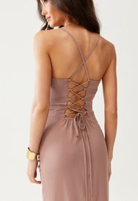 Žena oblečená v mauve šatech s křížkovým šněrováním na odhaleném zádech a tenkými ramínky, pózuje proti jednoduchému světlému pozadí.