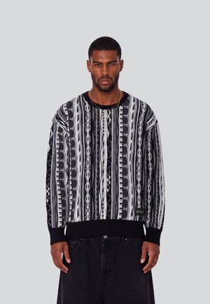 Mann mit kurzen Haaren und Bart, der einen schwarz-weiß-grau gemusterten Pullover und schwarze Hose trägt, steht vor einem einfarbigen grauen Hintergrund.