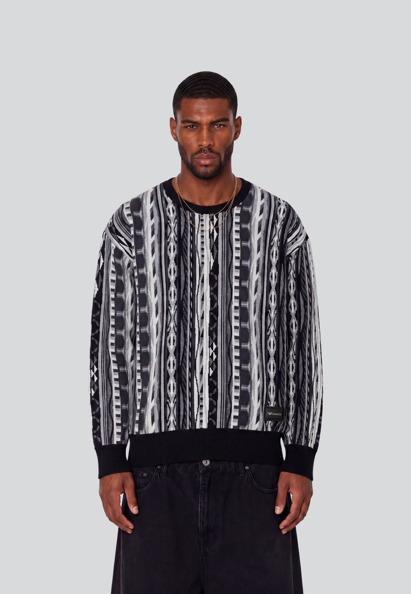 Homme aux cheveux courts et à la barbe, vêtu d’un pull à motifs noirs, blancs et gris et d’un pantalon noir, debout devant un fond gris uni.