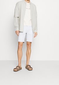 Muž v světle šedé košili na knoflíky, kterou má otevřenou přes bílý tričko s logem, světle modrých šortkách a hnědých sandálech se dvěma popruhy, stojící na jednobarevném pozadí.