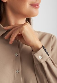 Beez blouse nupuga nööridega, pehme tekstuuri ja õrna sära ning musta etiketi ja irideerivate nööpidega.
