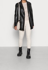 Blazer negro de cuero de doble botonadura sobre un suéter ligero de punto, combinado con jeans ajustados beige y botas negras con suela gruesa. Bolso bandolera claro.