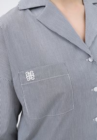 Randig skjorta med knäppning som har krage, vita knappar och en bröstficka med broderade detaljer. Tyget verkar lätt och mjukt.