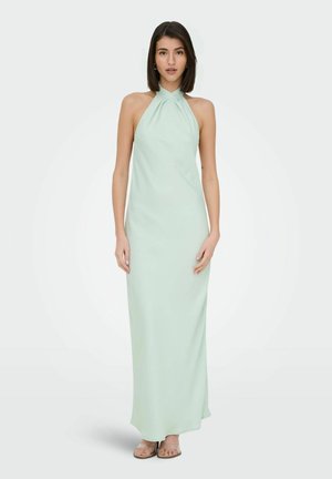 Femme debout portant une robe maxi verte menthe sans manches à col licou avec fines bretelles et sandales beige sur fond blanc.