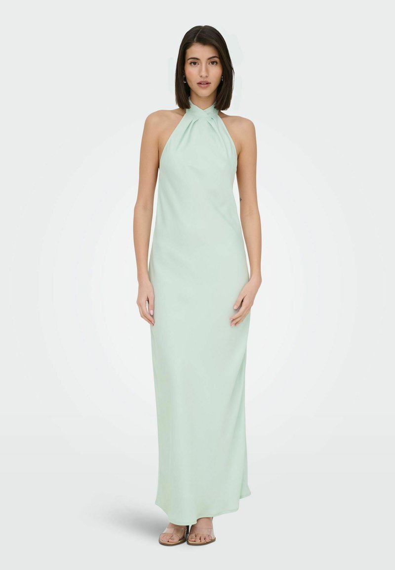Femme debout portant une robe maxi verte menthe sans manches à col licou avec fines bretelles et sandales beige sur fond blanc.